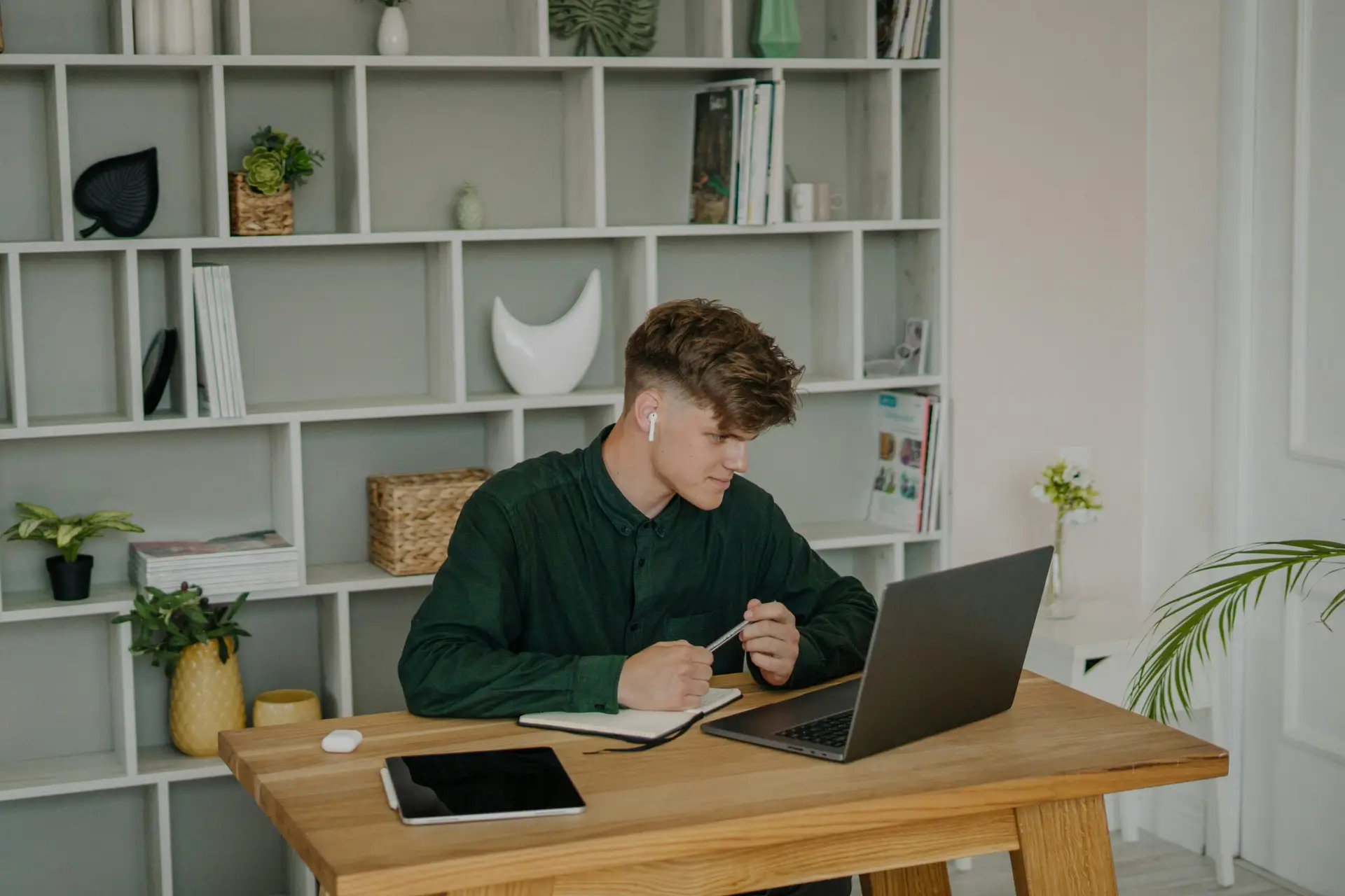 A young man using a laptop for online learning at home, seated at a wooden desk.